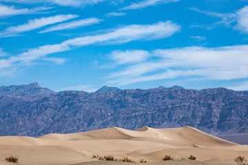 Fototapeta na wymiar Sand dunes in the foreground in the desert of Death Valley National park, in contrast with mountains in the background and a blue soft sky with moving clouds