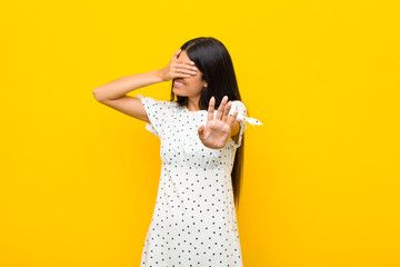 young pretty latin woman covering face with hand and putting other hand up front to stop camera,...