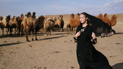beautiful woman national eastern clothes black color,veil,hijab,abaya ornament her head,portrait steppes camels,strong wind,desert,wildlife,well,decoration,caucasian,slow ,sun