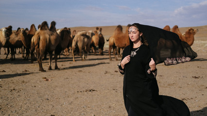 beautiful woman national eastern clothes black color,veil,hijab,abaya ornament her head,portrait steppes camels,strong wind,desert,wildlife,well,decoration,caucasian,slow ,sun
