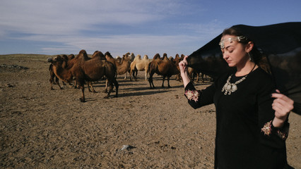 beautiful woman national eastern clothes black color,veil,hijab,abaya ornament her head,portrait steppes camels,strong wind,desert,wildlife,well,decoration,caucasian,slow ,sun