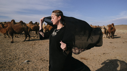 beautiful woman national eastern clothes black color,veil,hijab,abaya ornament her head,portrait steppes camels,strong wind,desert,wildlife,well,decoration,caucasian,slow ,sun