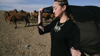 beautiful woman national eastern clothes black color,veil,hijab,abaya ornament her head,portrait steppes camels,strong wind,desert,wildlife,well,decoration,caucasian,slow ,sun