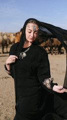 beautiful woman national eastern clothes black color,veil,hijab,abaya ornament her head,portrait steppes camels,strong wind,desert,wildlife,well,decoration,caucasian,slow ,sun