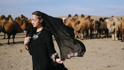 beautiful woman national eastern clothes black color,veil,hijab,abaya ornament her head,portrait steppes camels,strong wind,desert,wildlife,well,decoration,caucasian,slow ,sun