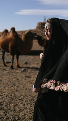 beautiful woman national eastern clothes black color,veil,hijab,abaya ornament her head,portrait steppes camels,strong wind,desert,wildlife,well,decoration,caucasian,slow ,sun