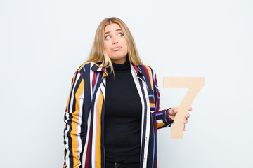 young pretty blonde woman confused, doubtful, thinking, holding a number 7.