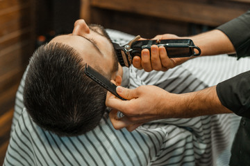 Beauty shop for men. Shaving a beard in a barbershop. Barber cuts his beard with a razor and clipper. close up Brutal haircuts. Hairdresser equipment. Selective focus.