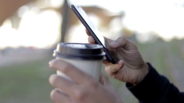 Cropped shot of man with coffee to go using smartphone. Close-up partial view of man texting via smartphone and drinking from paper cup, selective focus. Connection concept