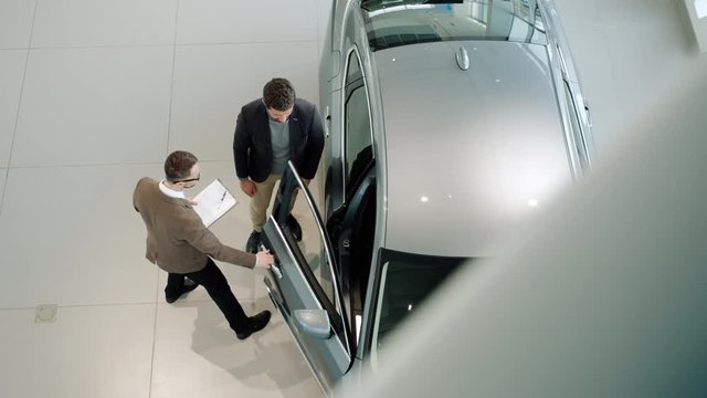 Sales manager is opening car door and inviting male customer to see the auto inside while buyer is choosing automobile in dealership. People and business concept.