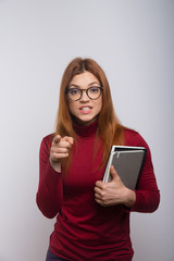 Naklejka premium Angry female student pointing at camera. Irritated young woman in eyeglasses holding books, pointing with finger and looking at camera. Education concept
