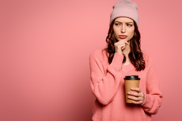 thoughtful girl looking away and touching chin while holding cofee to go on pink background