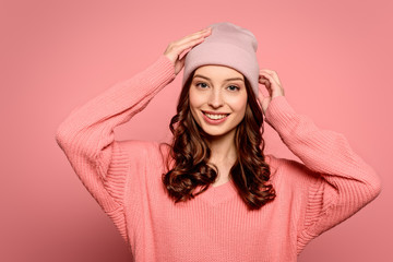 Obraz premium happy girl touching hat while smiling at camera on pink background