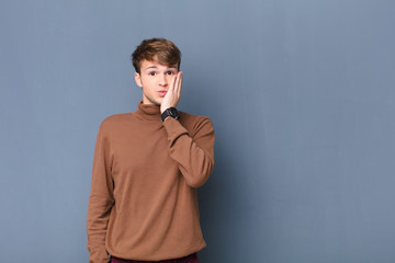 young blonde man feeling shocked and astonished holding face to hand in disbelief with mouth wide...