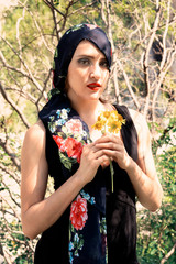 Beautiful young woman wearing head black scarf holding flower standing under the trees with sunlight, Mexico