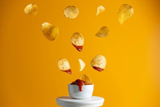 Tasty potato chips falling into blow with tomato sauce, frozen in the air. Yellow food art background.