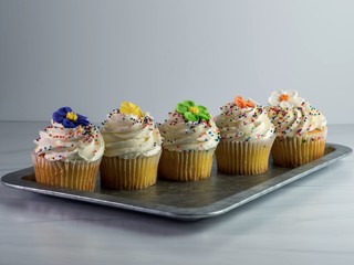 Rustic metal tray with 5 yellow cupcakes lined up decorated with white frosting, sprinkles and fondant flowers in purple, yellow, green, orange to celebrate a birthday or special occasion.