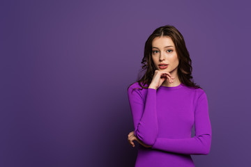 thoughtful girl holding hand near chin while looking at camera on purple background