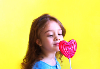 Large red heart - shaped candy is placed on the background of a cute little girl. Charming young lady looks at the lollipop with temptation. Spring or summer card with copy space. Focus on the candy