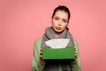 Fototapeta premium displeased, diseased girl in warm scarf taking box of paper napkins isolated on pink