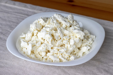 A white square plate filled with cottage cheese stands on a linen tablecloth on a wooden table. Selective focus