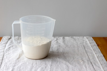 Plastic measuring cup with flour stands on a linen tablecloth on a wooden table against a gray...