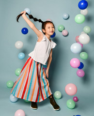Fototapeta premium Playful asian kid girl celebrates has fun surrounded by colorful air balloons holding up her long black plait