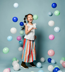 Fototapeta premium Laughing asian kid girl in party clothes is having fun surrounded by colorful air balloons holds hand up with open palm