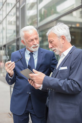 Naklejka na ściany i meble Two senior caucasian businessmen talking in front of an office building, working on a tablet and discussing