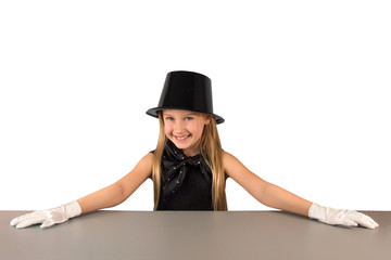 Cheerful charming little eight-year-old magician girl in hat and gloves is preparing to show tricks and presentations at gray table on white background. Concept of discounts for children's products