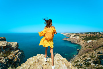 Rear view of traveler woman standing on top rock beach with backpack, bright yellow dress and hair blowing in the wind.