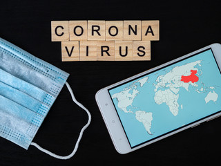 Coronavirus word made of wood block, world map on smartphone screen and breathing mask. Coronavirus text on dramatic atmosphere black wooden table. Coronavirus concept top view.