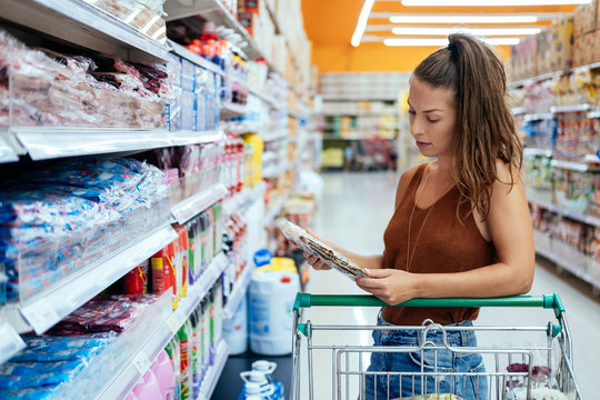 Woman Shopping In Supermarket Reading Product Information Stock Photo. Young Smiling Woman Groceries Shopping In Local Supermarket. She Is Standing,holding Product And Reading Nutrition Facts.