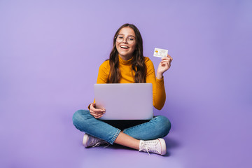 Image of young woman holding credit card and using laptop while sitting