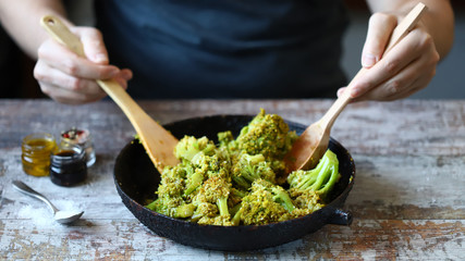 A chef with a wooden spatula and spoon mixes broccoli in a pan.