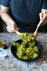 A chef with a wooden spatula and spoon mixes broccoli in a pan.