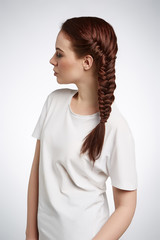 A young red-haired girl with a graceful French braid. The lady in an ivory t-shirt is posing sideways on the gray background and looking away. 