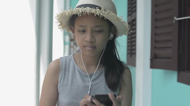 Asian cute girl in casual dress wearing straw hat using mobile phone and earphones walking leisurely in side arcade in chino portuguese style building, Phuket, Thailand. Slow motion.