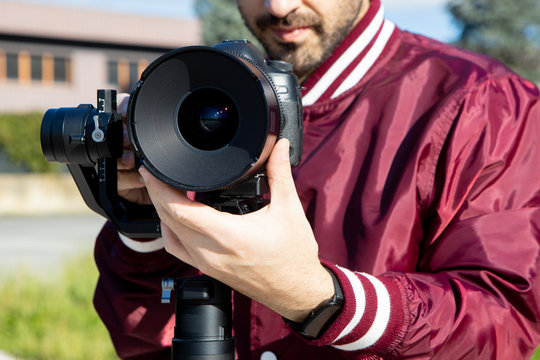 Caucasian young man with reverse cap and red jacket works outdoor with his camera on a gimbal stabiliser. Video operator focusing with his video camera. Cinema lens on DLSR camera. Close up angle shot