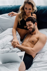 Smiling woman in bra looking at laptop near handsome boyfriend on bed