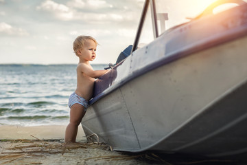 Cute adorable caucasian blond little toddler boy in white pants standing on sandy beach near fishing boat look and dreaming about sailor profession in future. Travel and adventure concept.Summer day