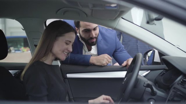 Young handsome Middle Eastern man talking with beautiful Caucasian woman sitting on driver's seat. Successful businessman buying automobile for girlfriend or wife in dealership. Car industry, joy.