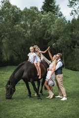 Loving family, father, mother and two little daughters riding horse at countryside outdoors. Young happy family having fun, giving high five to each other, family time concept. Summertime.