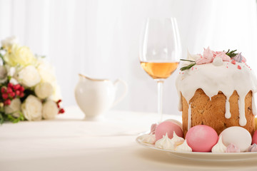 Selective focus of delicious Easter cake decorated with meringue with pink and white eggs on plate near wine glass and flowers