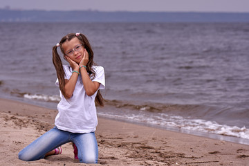Portrait of a teenage girl in glasses and a white T-shirt walking on a sandy shore on a cloudy...