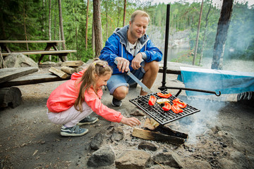 Man and his little daughter having barbecue in forest on rocky shore of lake, making a fire,...