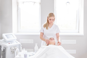 Pretty young woman beautician does antiaging treatments on face of young pretty female client lying on couch under white blanket in modern high-tech spa salon. Concept of modern cosmetology procedures