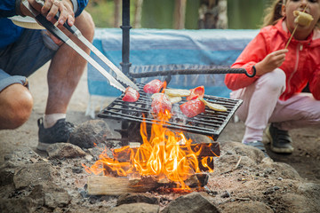 Man and his little daughter having barbecue in forest on rocky shore of lake, making a fire, grilling bread, vegetables and marshmallow. Family exploring Finland. Scandinavian summer landscape. 