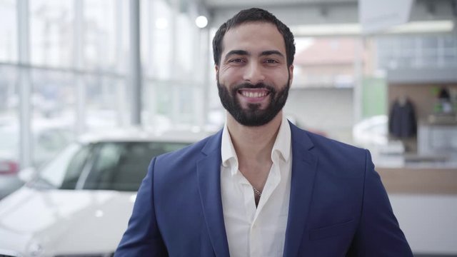 Portrait of successful Middle Eastern man posing in car dealership. Handsome young businessman looking back at new white vehicle, turning to camera and smiling. Success, lifestyle, business.