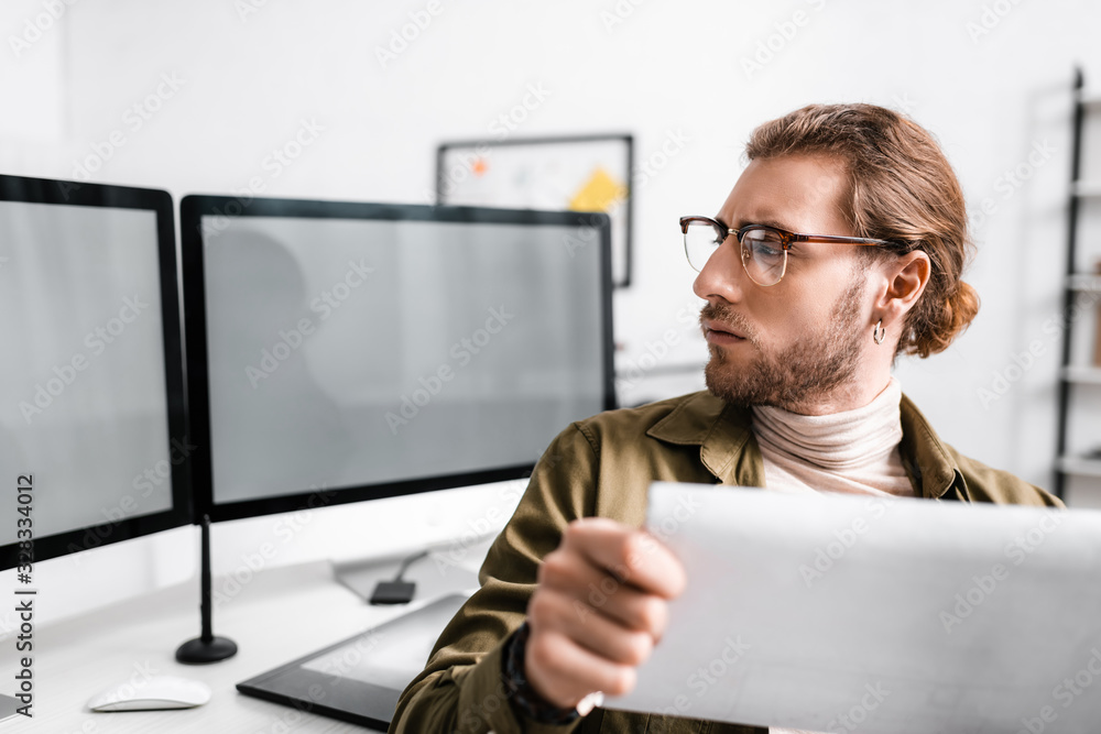 Poster selective focus of handsome digital designer holding blueprint and looking at computer monitors on t